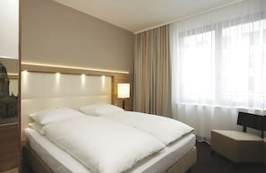 Habitación de hotel con una cama grande, una mesita de noche, una lámpara y vistas de la ciudad a través de cortinas translúcidas.