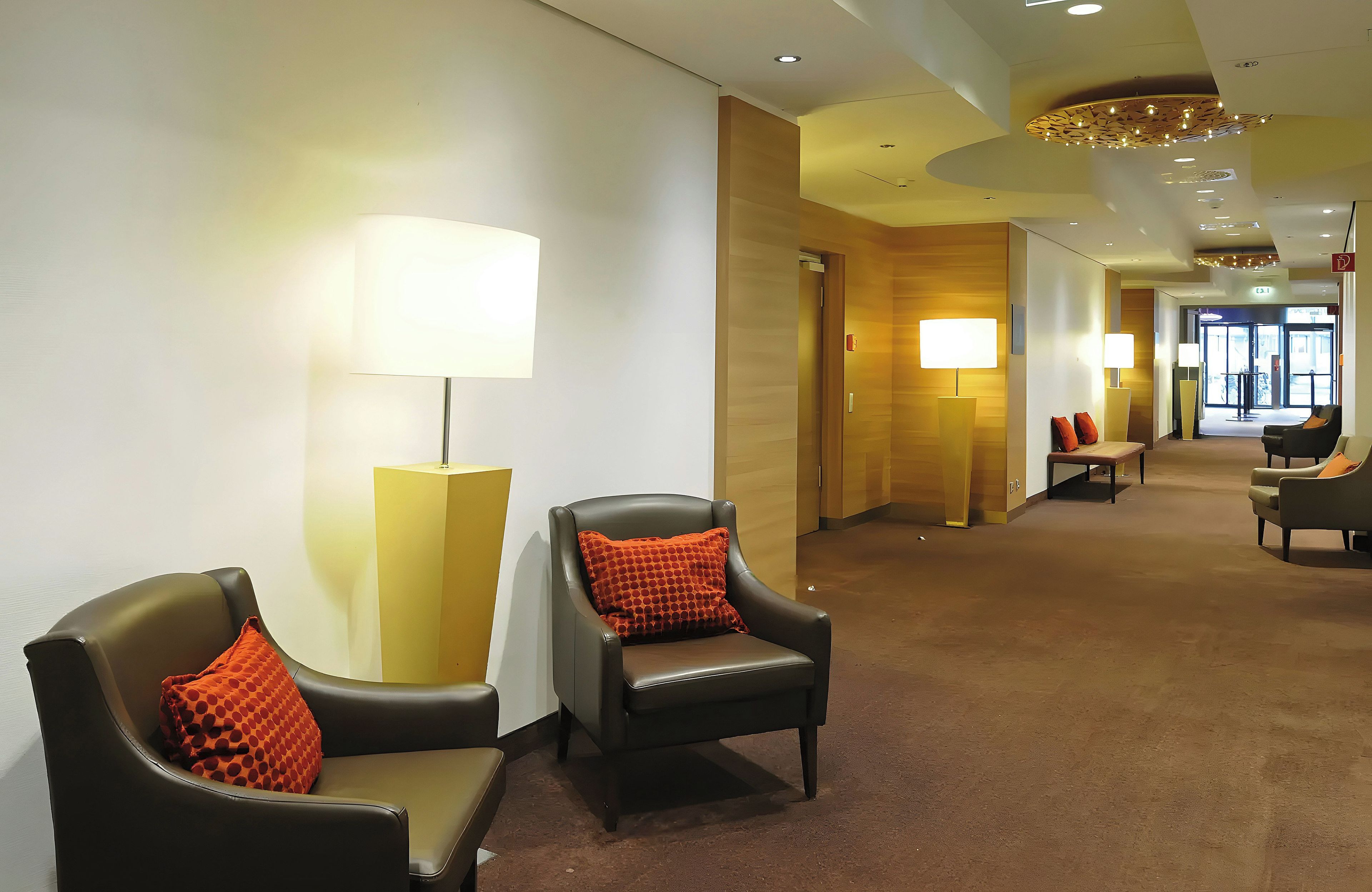 Una hall d'albergo con zone salotto, lampade e un corridoio che porta a un'uscita.