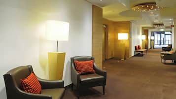 Una hall d'albergo con zone salotto, lampade e un corridoio che porta a un'uscita.