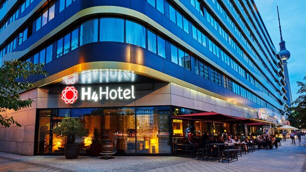 Ein modernes Hotel mit einem gut sichtbaren H4 Hotel Schild, Außenbereich mit Sitzgelegenheiten und Blick auf die Skyline der Stadt.