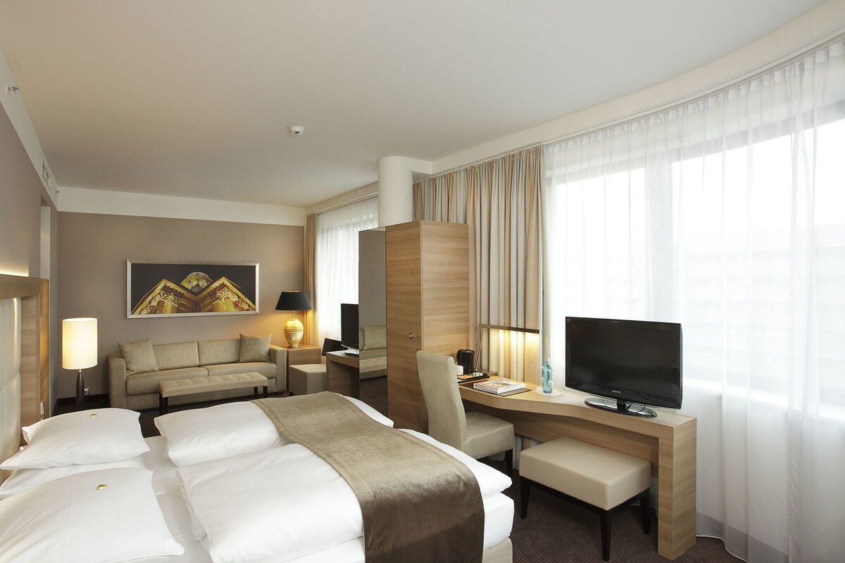 A hotel room with a bed, desk, TV, and a large window with curtains.