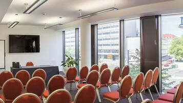 Un salón de conferencias con filas de sillas, un ventanal con vistas a edificios y un televisor de pantalla plana en la pared.