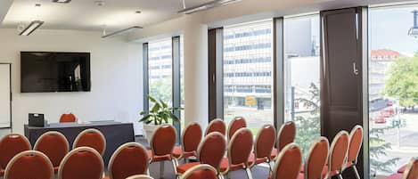 Une salle de conférence avec des rangées de chaises, une grande fenêtre donnant sur des immeubles et un téléviseur à écran plat fixé au mur.