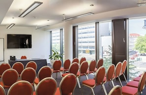 Un salón de conferencias con filas de sillas, un ventanal con vistas a edificios y un televisor de pantalla plana en la pared.