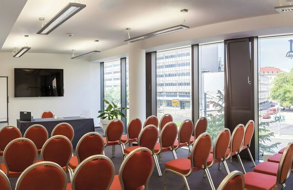 Un salón de conferencias con filas de sillas, un ventanal con vistas a edificios y un televisor de pantalla plana en la pared.