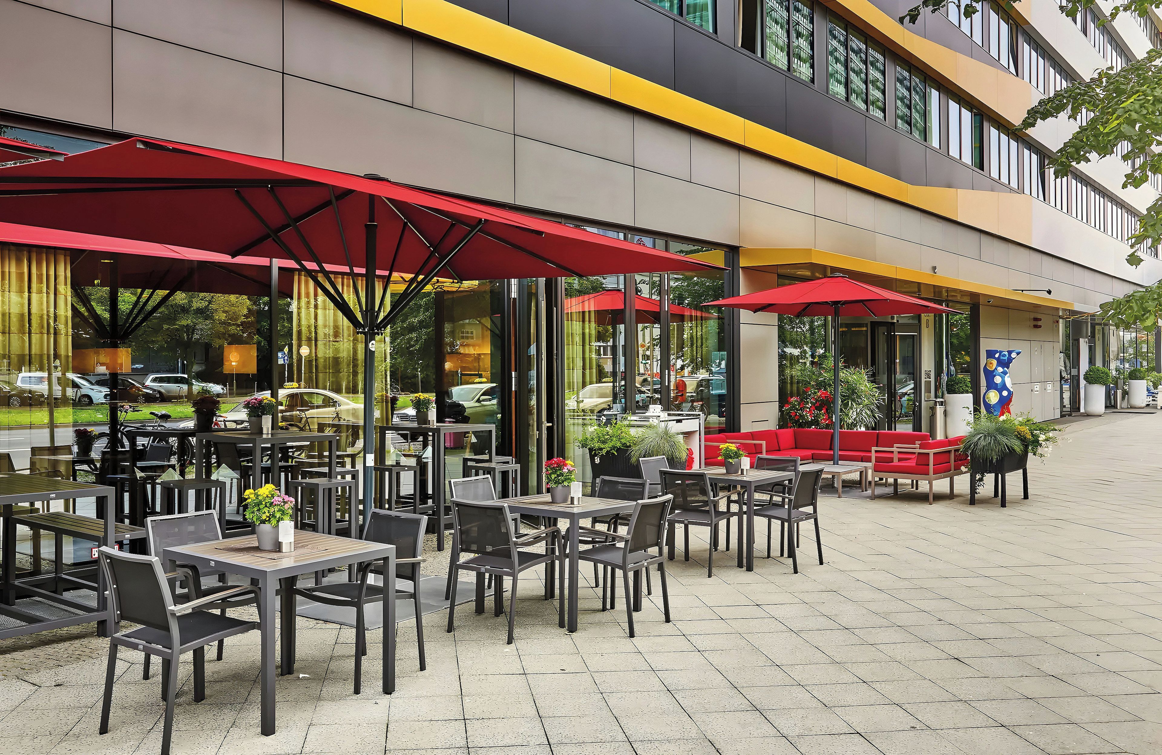 An outdoor cafe area with tables and chairs, red umbrellas, and potted plants.