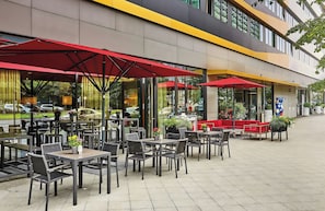 Un área de café al aire libre con mesas, sillas, sombrillas rojas y plantas en macetas.