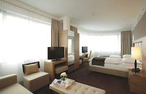 A hotel room with a bed, a desk with a computer, a television, a seating area with a sofa, and a window with curtains. - Hotel Berlin Alexanderplatz by Leonardo Hotels (Berlin)