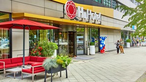 Ein modernes Hotelgebäude mit einem roten Sitzbereich im Freien, einem großen Kübelpflanzen und einer auffälligen, blau-gelben Skulptur.