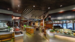 Ein modernes Restaurantambiente mit Holzelementen, einer zentralen Anrichte mit Speisenauslagen und einer Sitzgegend mit Tischen und Stühlen.