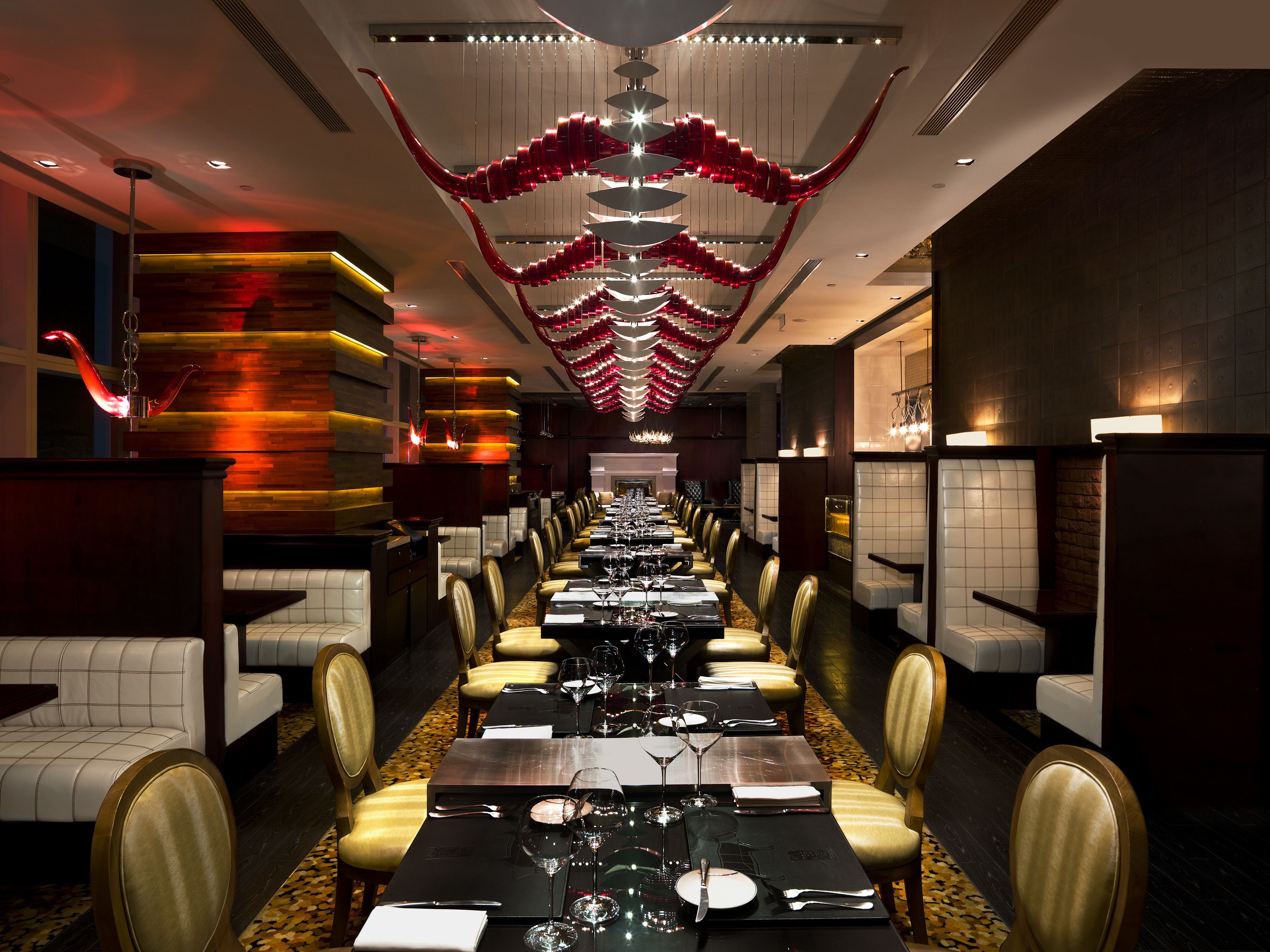 A long dining area with tables set for multiple guests, featuring a modern chandelier and a patterned wall design.