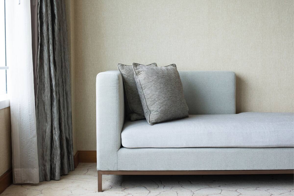 A light grey sofa with two grey cushions against a plain wall, a window with grey curtains, and a patterned carpet.