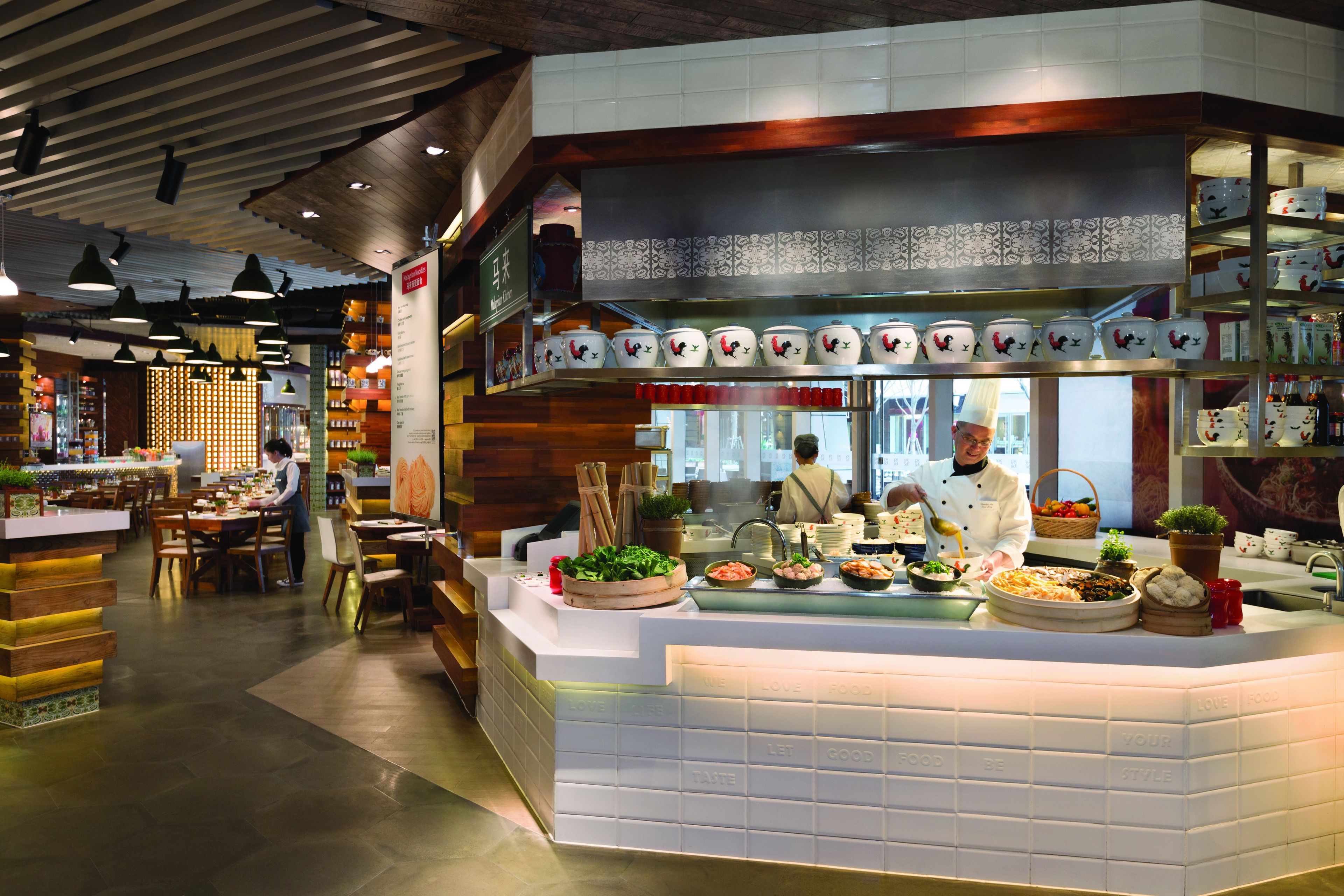 Un restaurante con una decoración moderna, que presenta una barra central donde un chef prepara comida, rodeada de diferentes platos y utensilios de cocina.