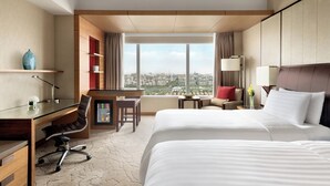 Ein Hotelzimmer mit zwei Betten, einem Schreibtisch, einem Sessel, einem kleinen Tisch und Blick auf die Stadt.