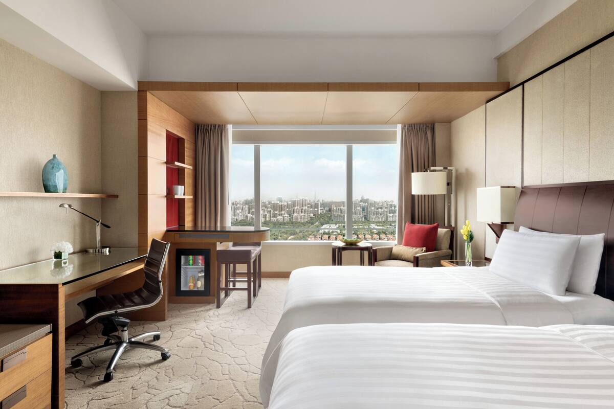 A hotel room with two beds, a desk, a chair, a small table, and a view of the city.