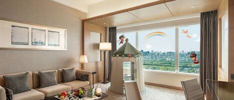 A hotel room with a sofa, a coffee table, a play area with a slide, and large windows offering a city view.