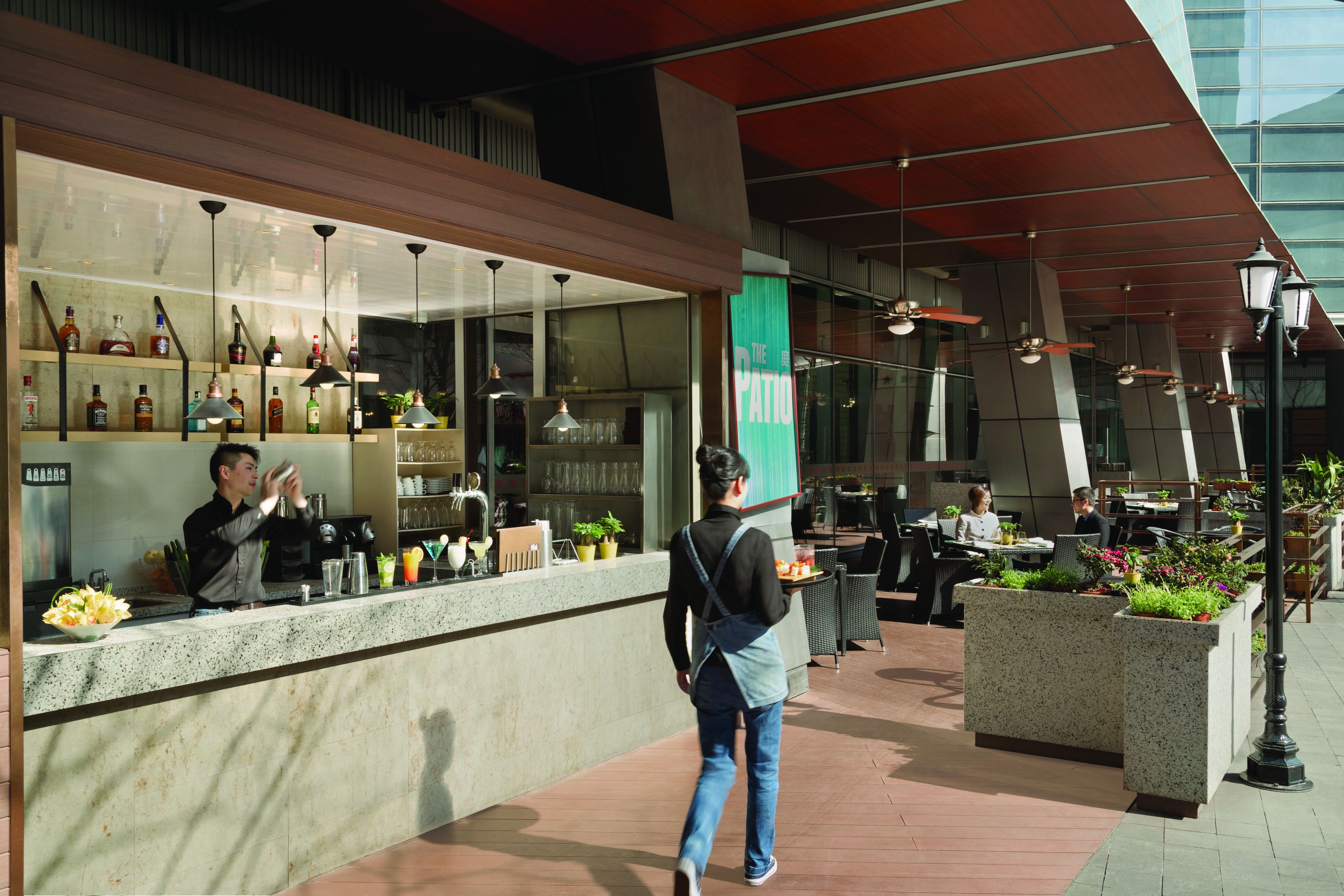 Un bar moderno con mostrador, taburetes y una variedad de botellas. Hay zonas de estar al aire libre con mesas y sillas.