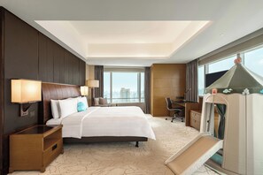 A hotel room with a large bed, a desk, a chair, a TV, and a view of the city. - Kerry Hotel Pudong Shanghai (Shanghai)