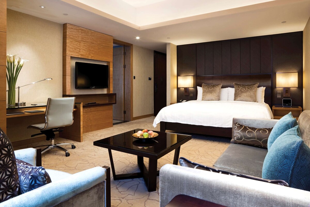 Hotelkamer met een bed, een bureau met stoel, een televisie, een bank en een salontafel met fruit.