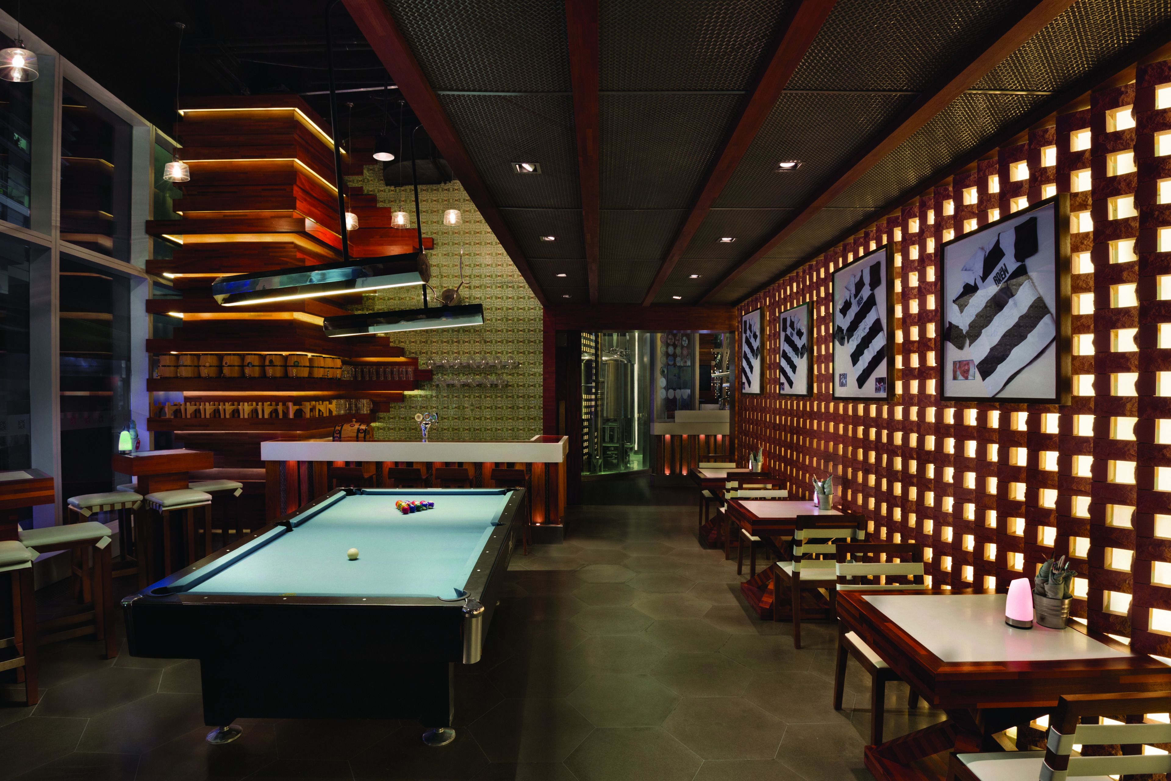 Une table de billard dans une pièce à la lumière tamisée, avec des meubles en bois et un mur en briques agrémenté de panneaux lumineux.