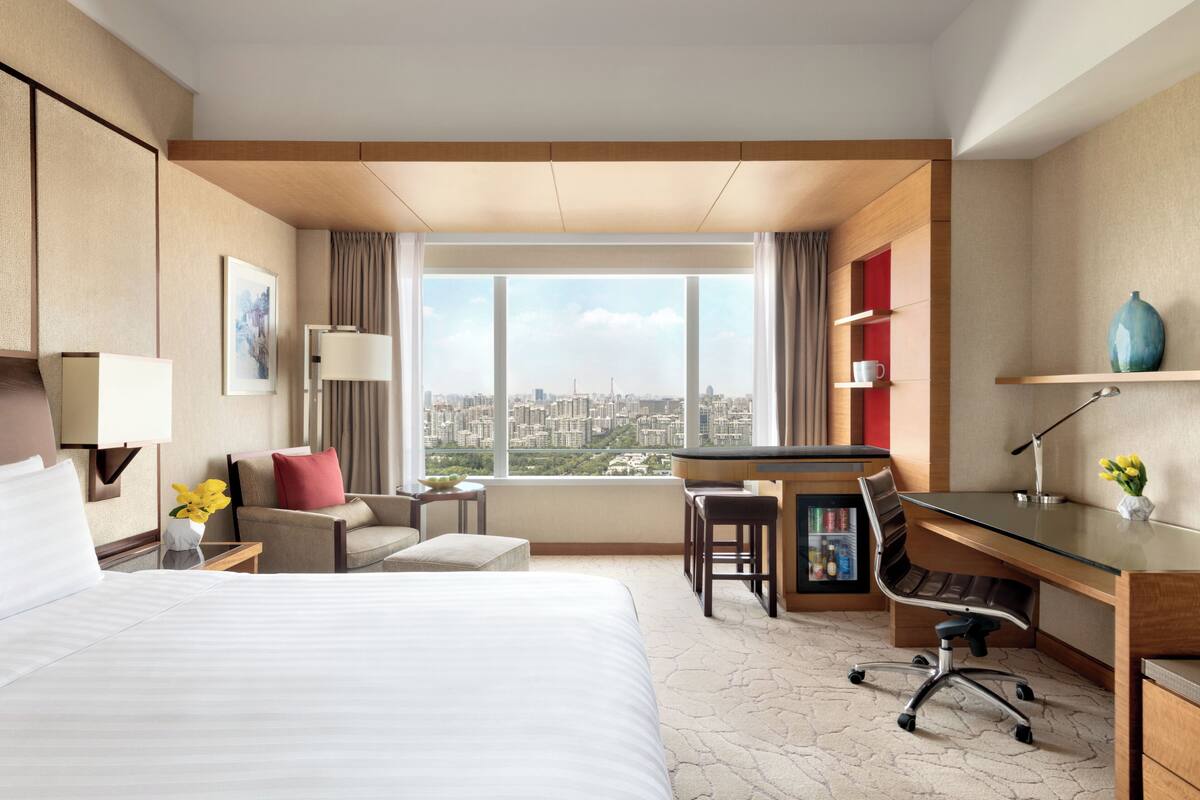 A hotel room with a bed, desk, and chair, a city view, and a minibar.