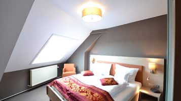 Un dormitorio con techo inclinado, tragaluz, cama con colcha estampada, mesita de noche con lámpara y mesita auxiliar.