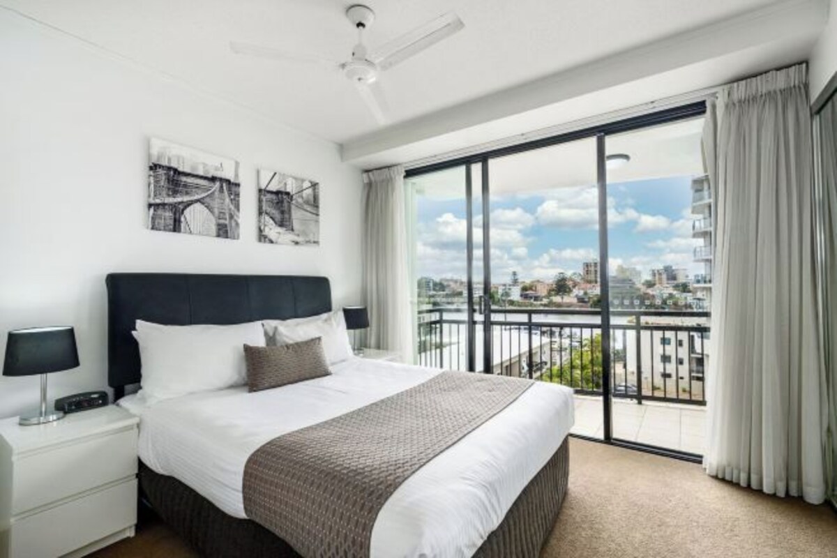 A hotel room with a large bed, a nightstand, a ceiling fan, and a view of the city through a large window.