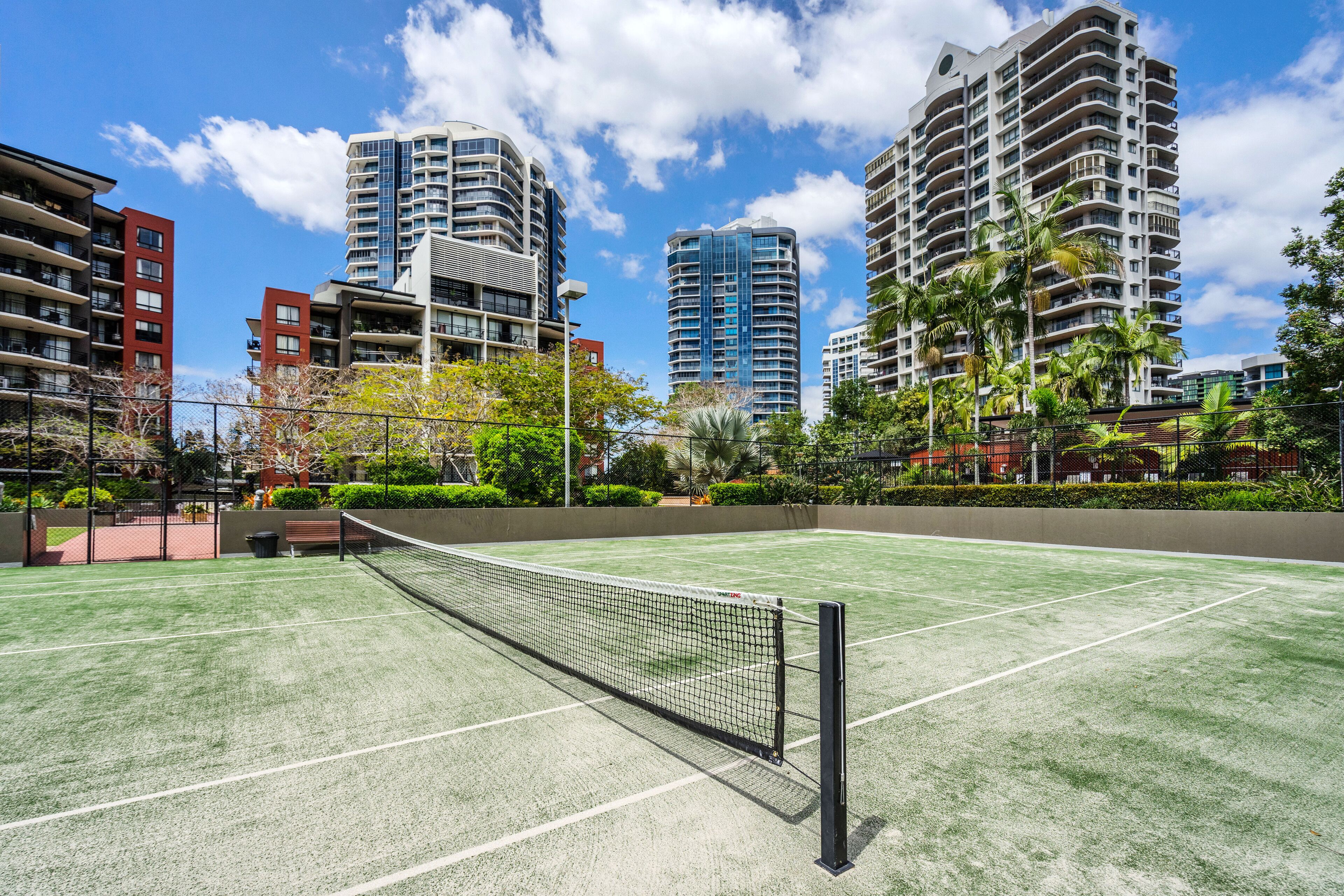 Un court de tennis en plein air, doté d’un filet, entouré de grands immeubles résidentiels et de verdure.