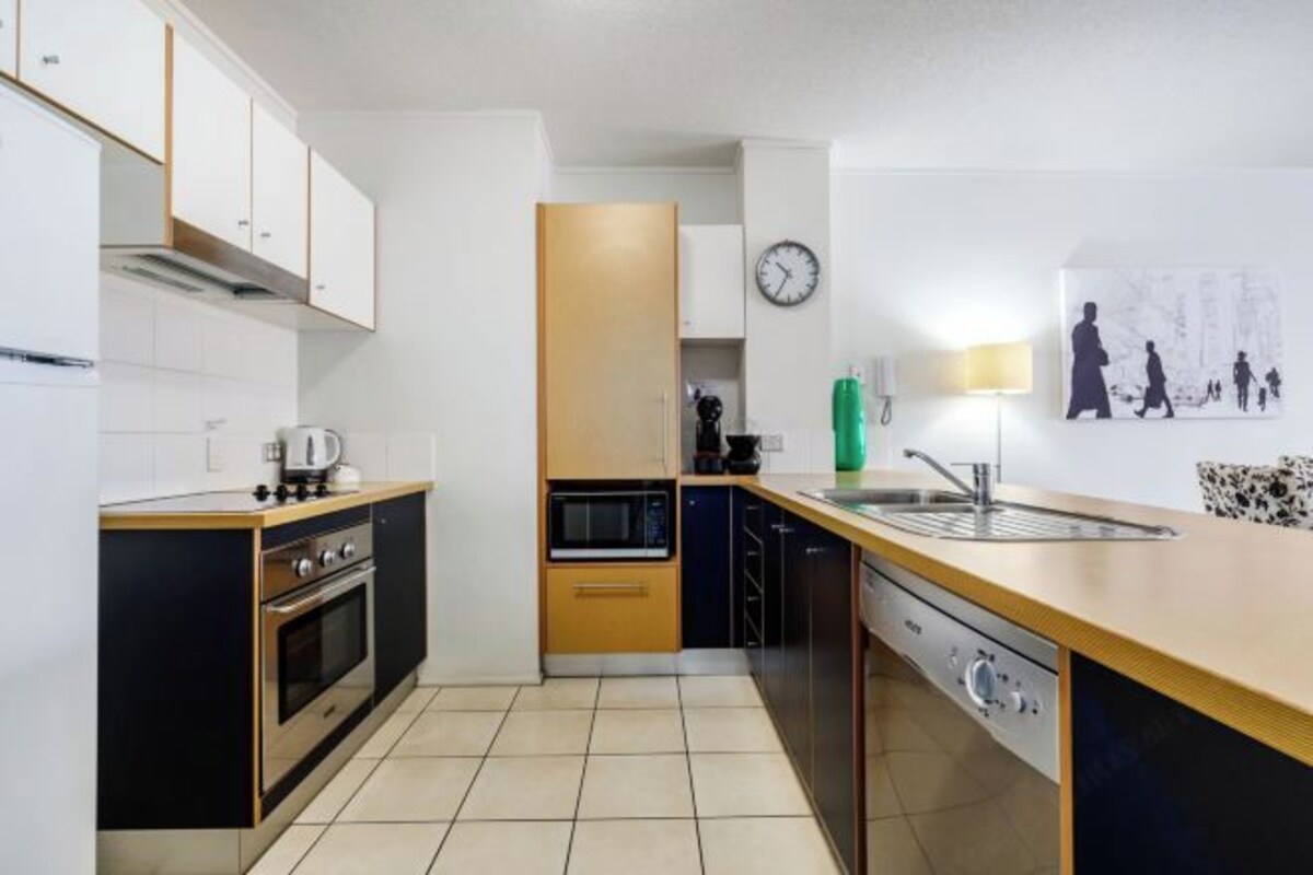 A modern kitchen with a central island, built-in oven, microwave, and dishwasher. There is a clock on the wall and a green water bottle on the counter.