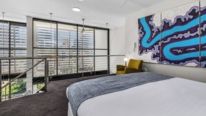 A hotel room with a large bed, a view of the city through floor-to-ceiling windows, a ceiling fan, and a modern abstract painting on the wall.