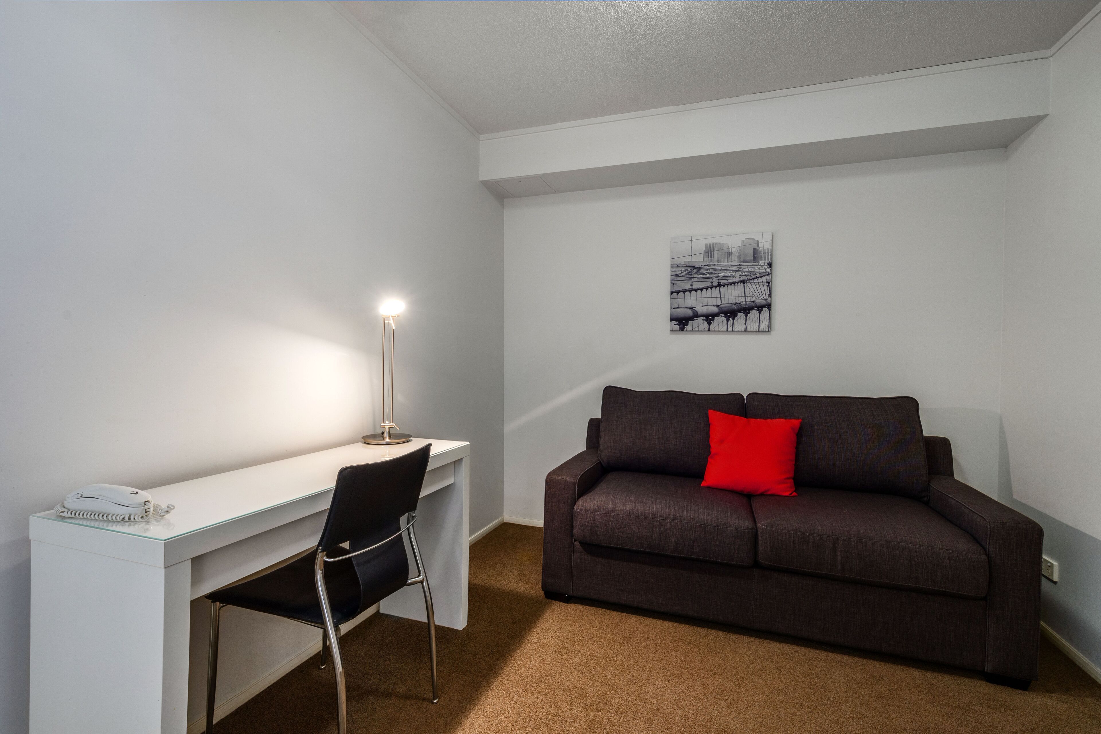 Une petite pièce moderne avec un bureau blanc, une chaise noire, un canapé marron agrémenté d’un coussin rouge et une œuvre d’art représentant un paysage urbain accrochée au mur.