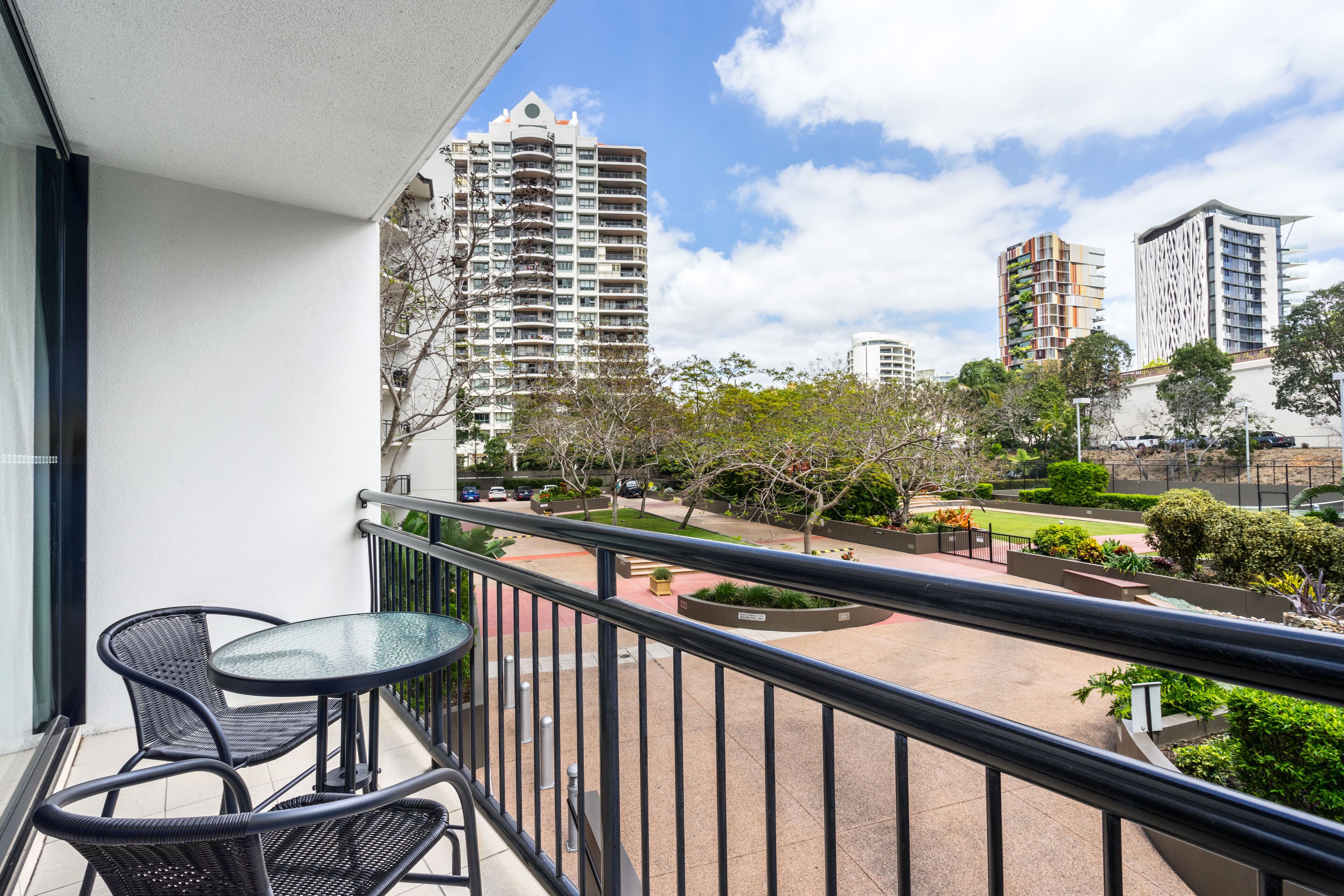 A balcony with two chairs and a small table, overlooking a landscaped area with trees and a road.