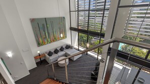 A modern interior with a staircase, a grey sofa, a colorful abstract painting, and a city view through large windows.