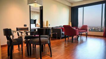 A modern hotel room with a dining area, a sofa, and a large window offering a city view.