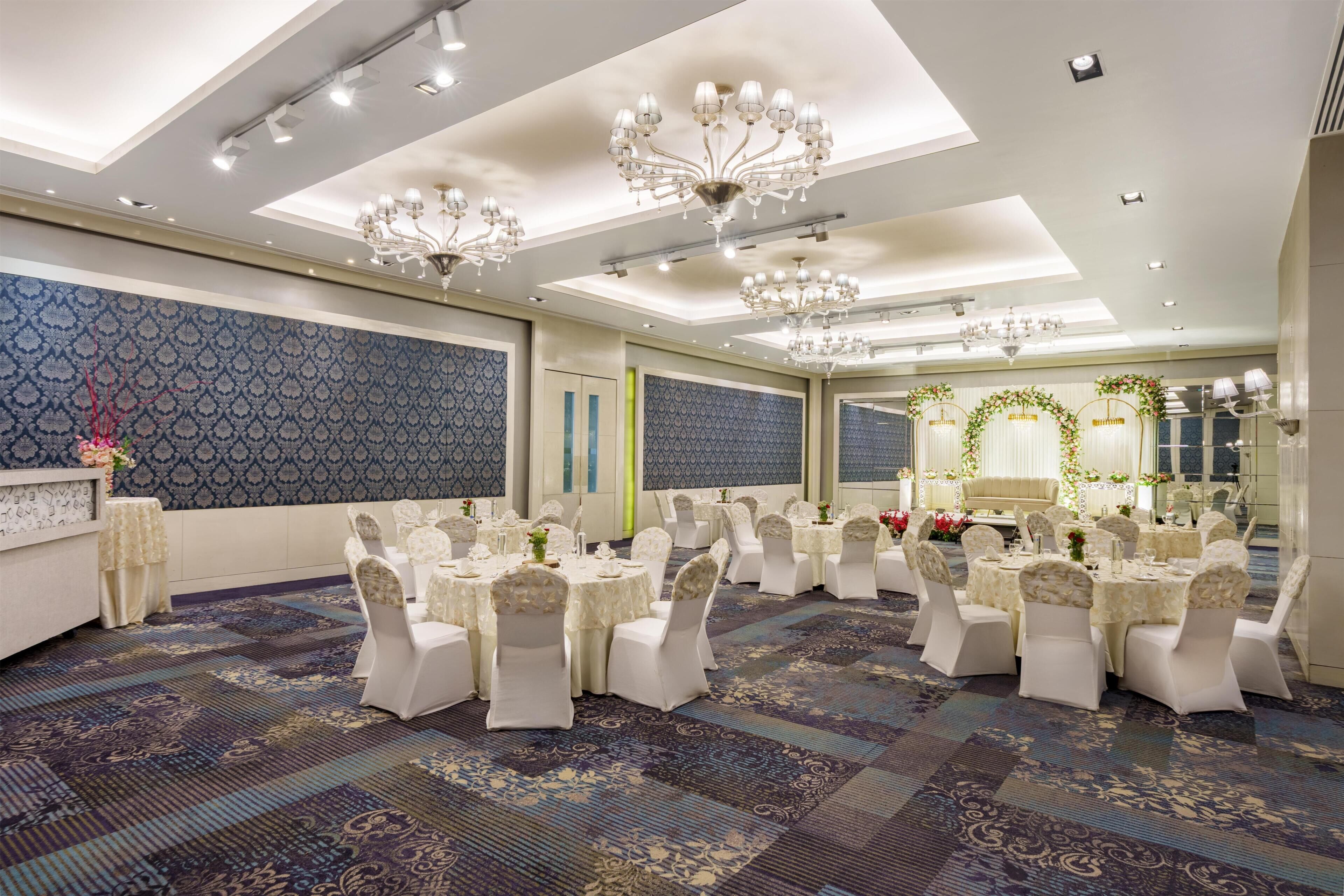 A banquet hall with round tables, white chairs, chandeliers, patterned carpet, and a large mirror.