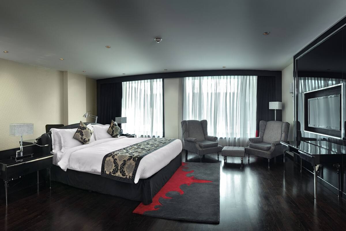 Une chambre d’hôtel moderne dotée d’un grand lit, d’un coin salon avec un fauteuil et un pouf, d’une télévision à écran plat et d’une grande fenêtre avec des rideaux.