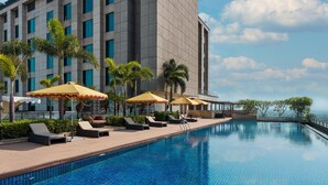 A swimming pool with lounge chairs and umbrellas, a large building in the background, and a clear sky.