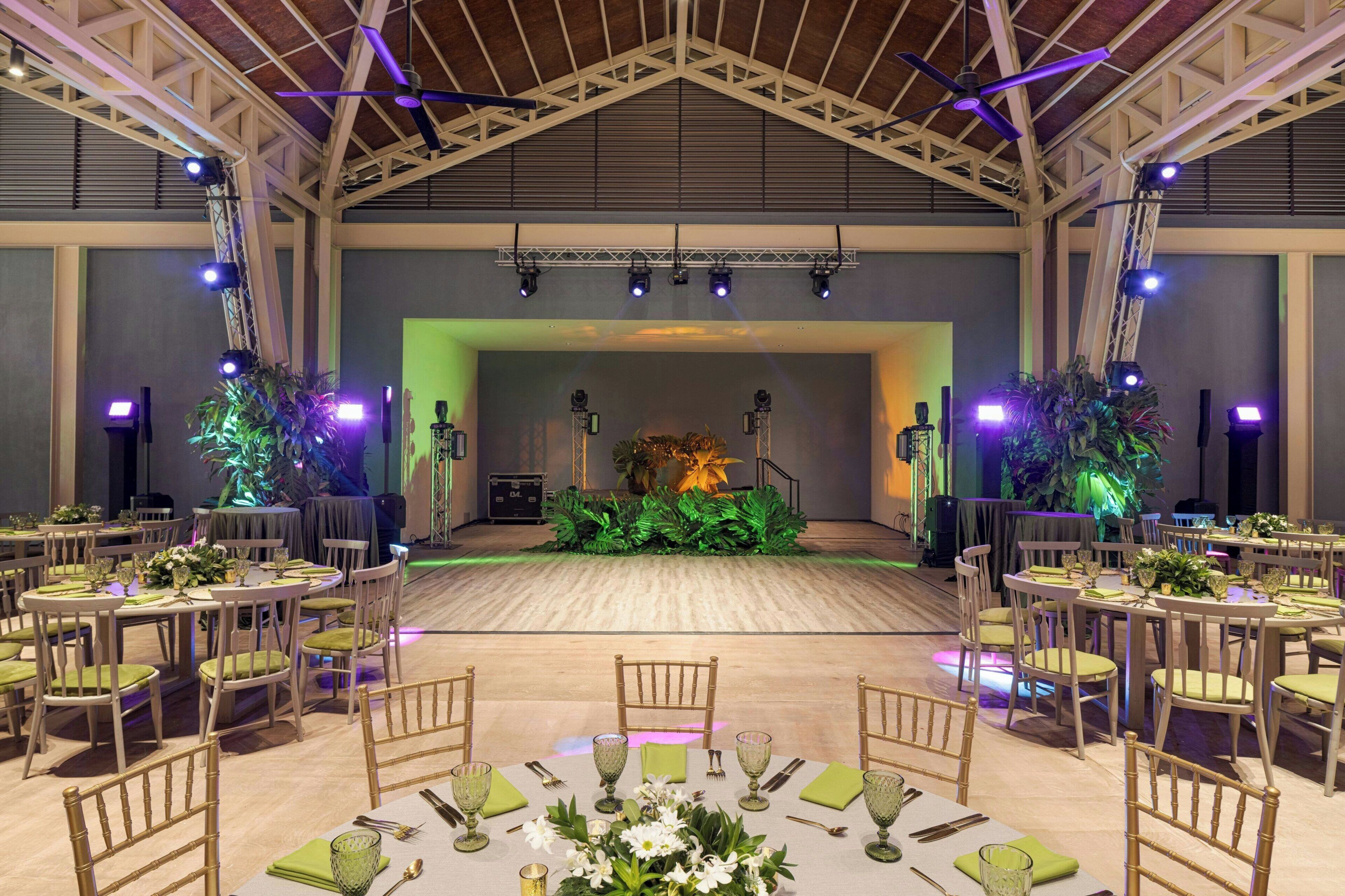 A spacious hall with a high ceiling, multiple tables set for dining, and a stage with lighting equipment.