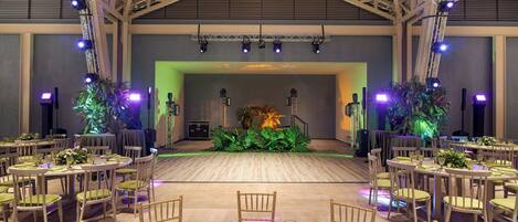A spacious hall with a high ceiling, multiple tables set for dining, and a stage with lighting equipment.