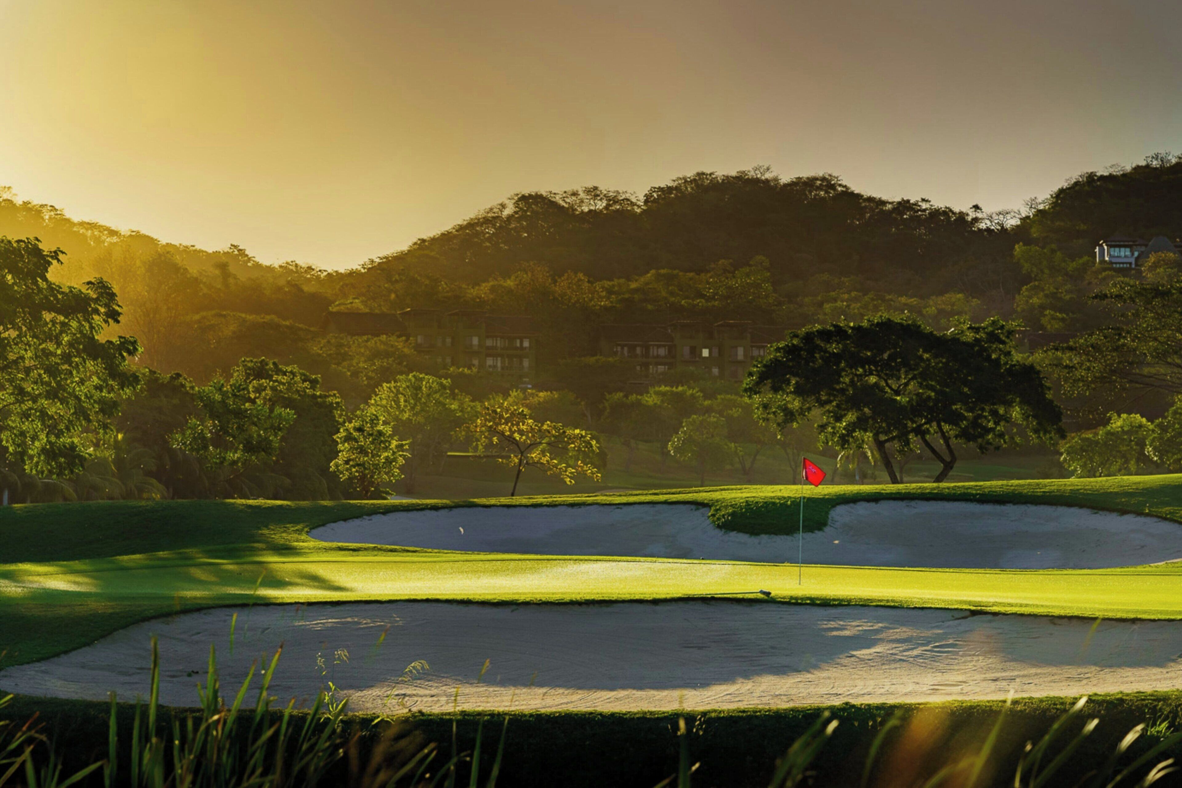 Campo de golf con búnkeres de arena, una bandera y un edificio al fondo.