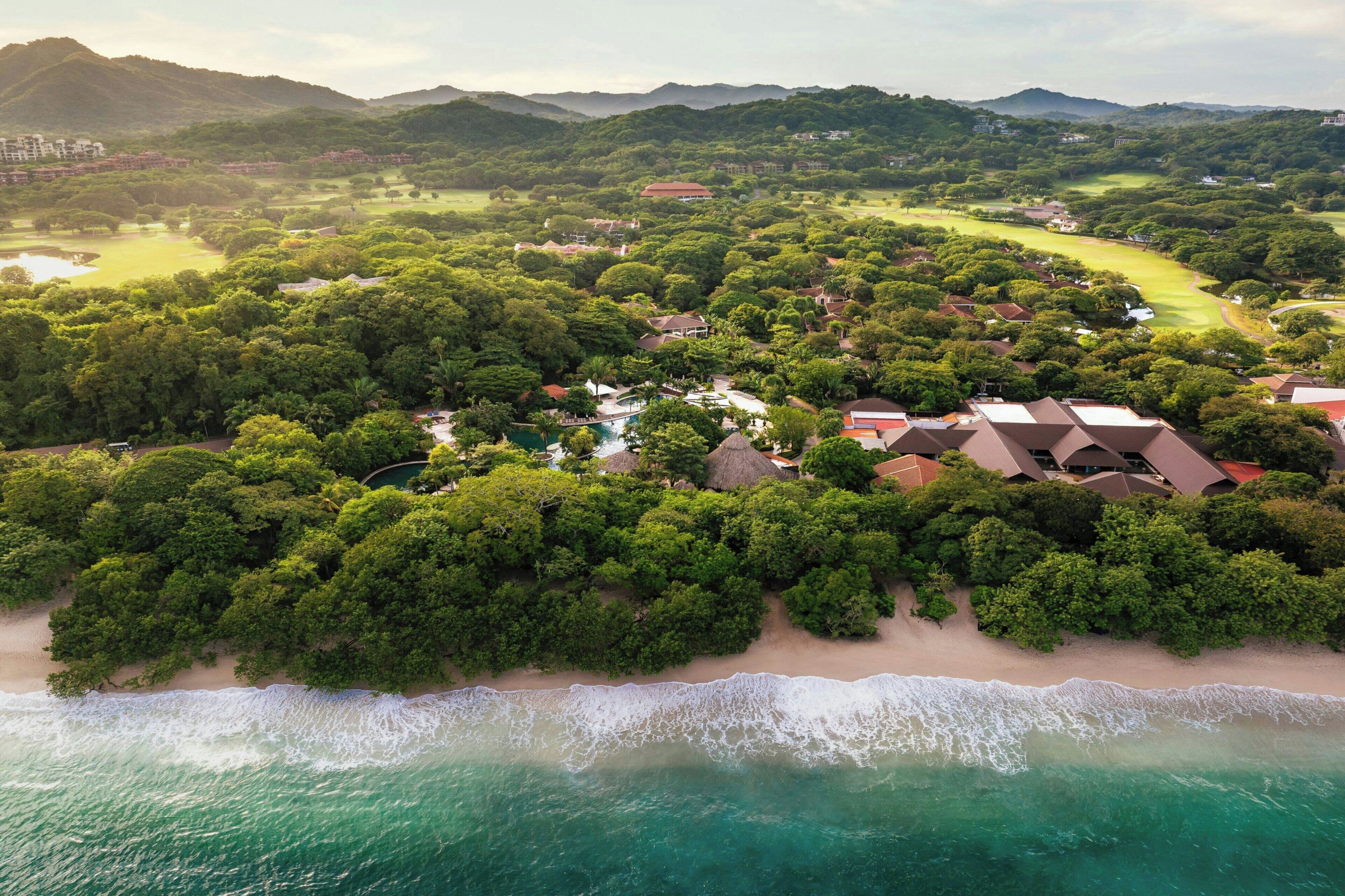 Vista aérea de un complejo turístico costero con playa, exuberante vegetación y un campo de golf.