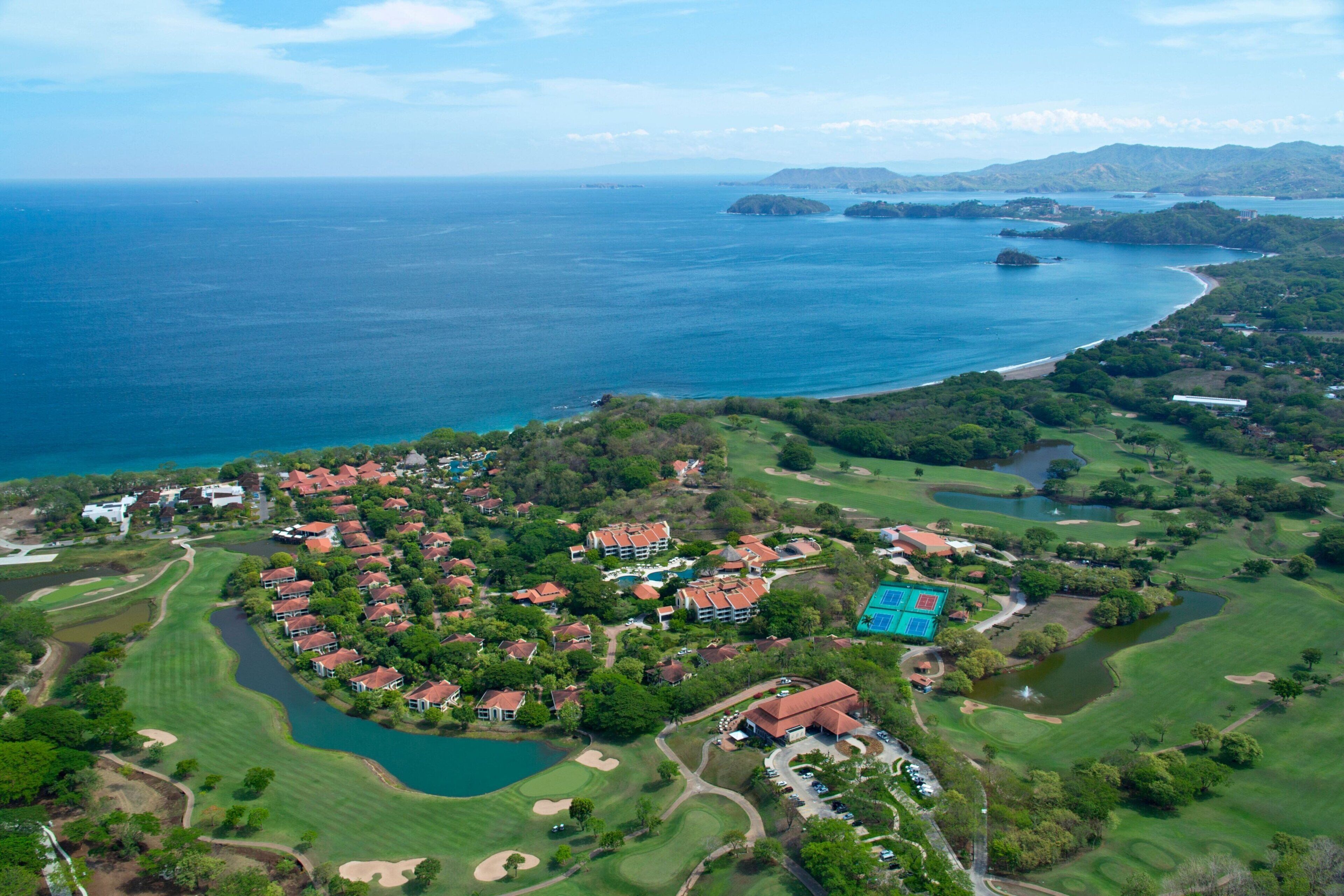 An aerial view of a coastal resort with multiple buildings, a golf course, and a large body of water.