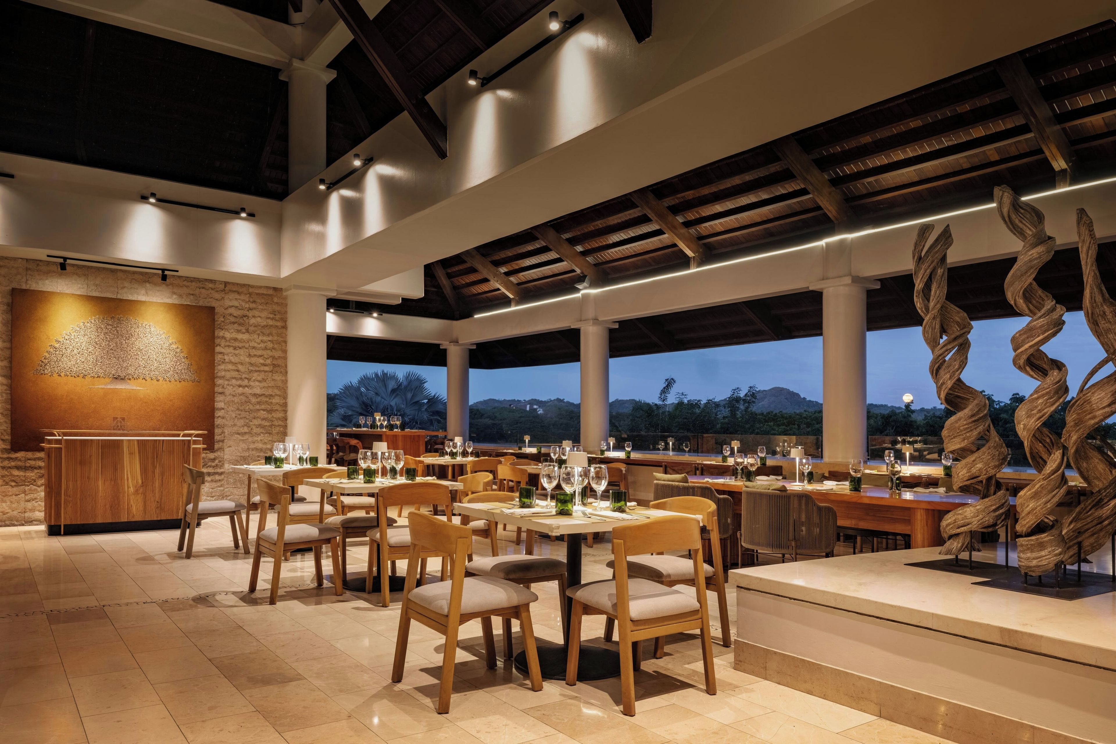 Een restaurant met meerdere tafels en stoelen, een groot houten sculptuur en uitzicht op het omliggende landschap.