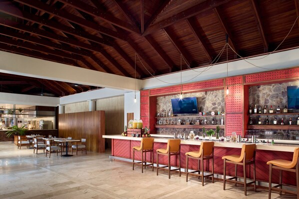 A bar area with wooden ceiling beams, a stone wall with a TV, and a bar counter with stools and bottles. - The Westin Reserva Conchal, an All-Inclusive Golf Resort & Spa (Cabo Velas)