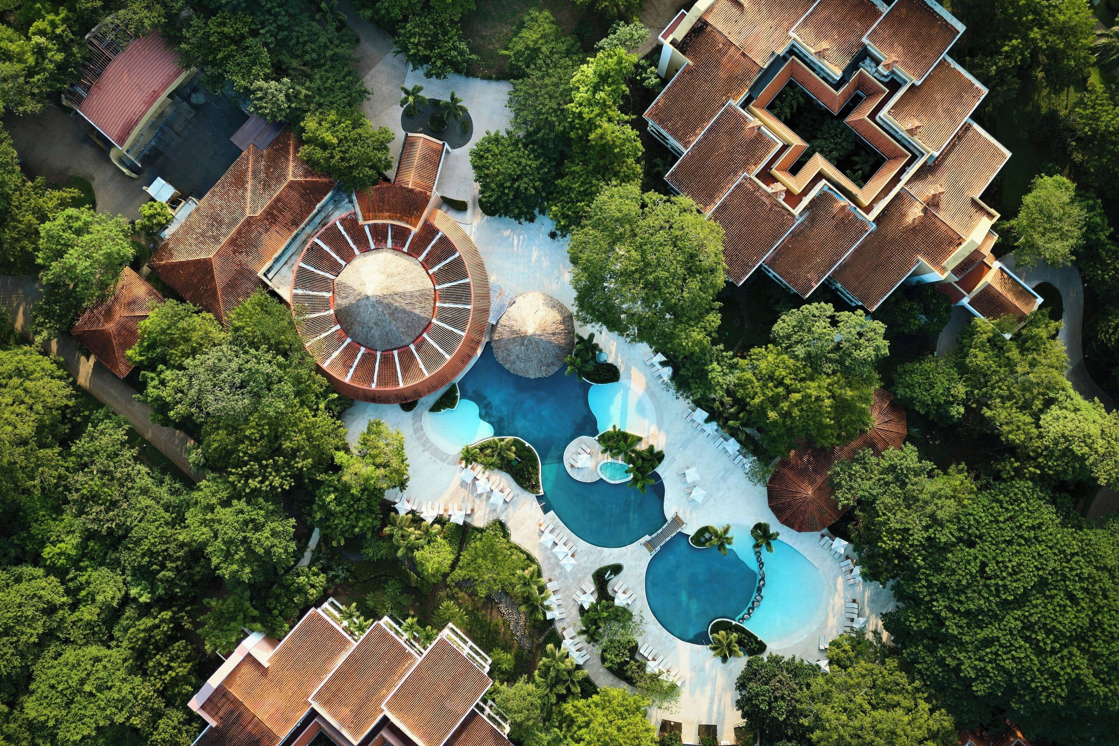 An aerial view of a resort with multiple swimming pools, a central circular water feature, and surrounding lush greenery.