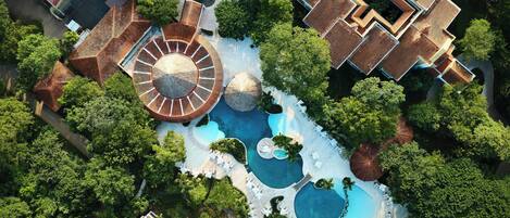An aerial view of a resort with multiple swimming pools, a central circular water feature, and surrounding lush greenery.