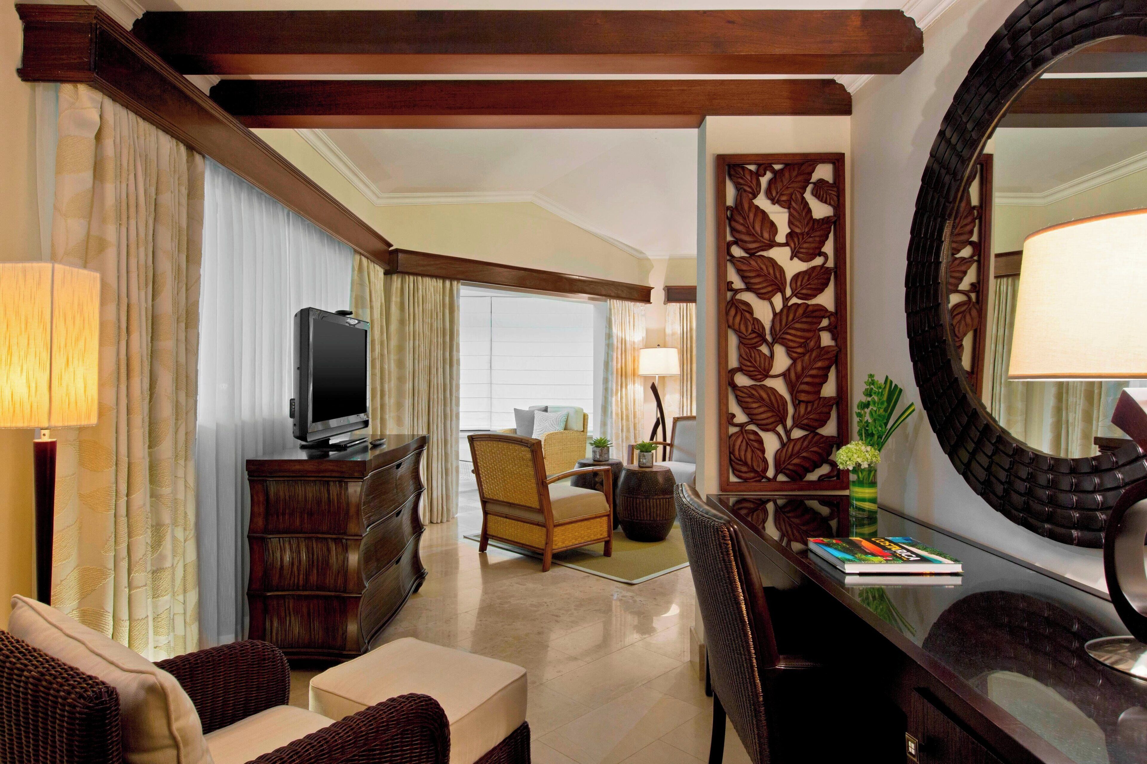 Habitación de hotel con televisión, sofá, silla, escritorio con espejo y un panel decorativo en la pared.
