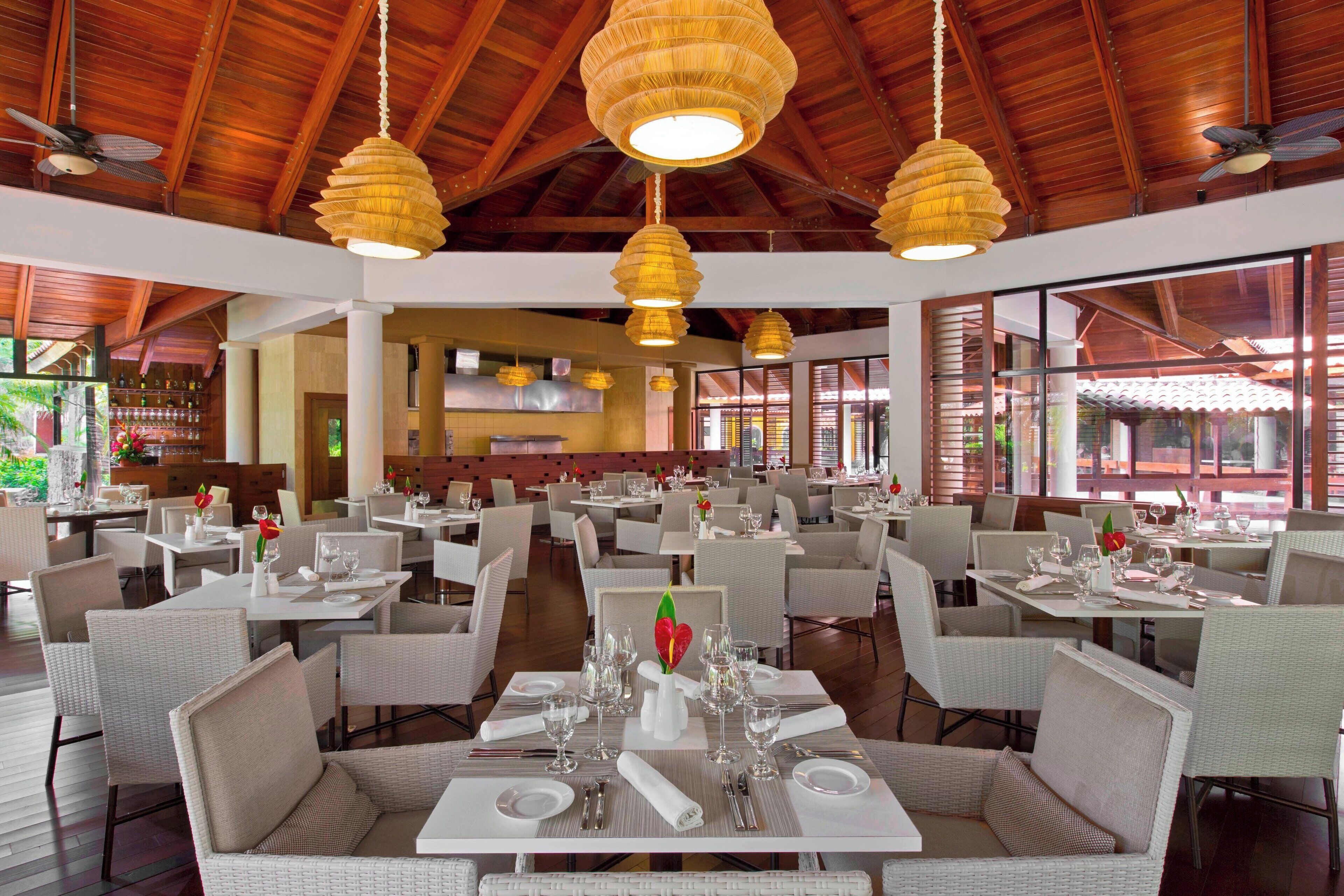 Uma ampla sala de jantar com várias mesas preparadas para refeições, com cadeiras de vime e um elemento decorativo central.