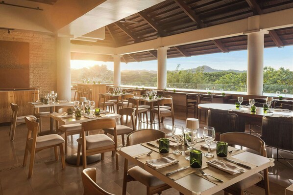 Un restaurante con mesas dispuestas para comer, amplias ventanas que ofrecen vistas al paisaje y un techo alto con vigas a la vista.