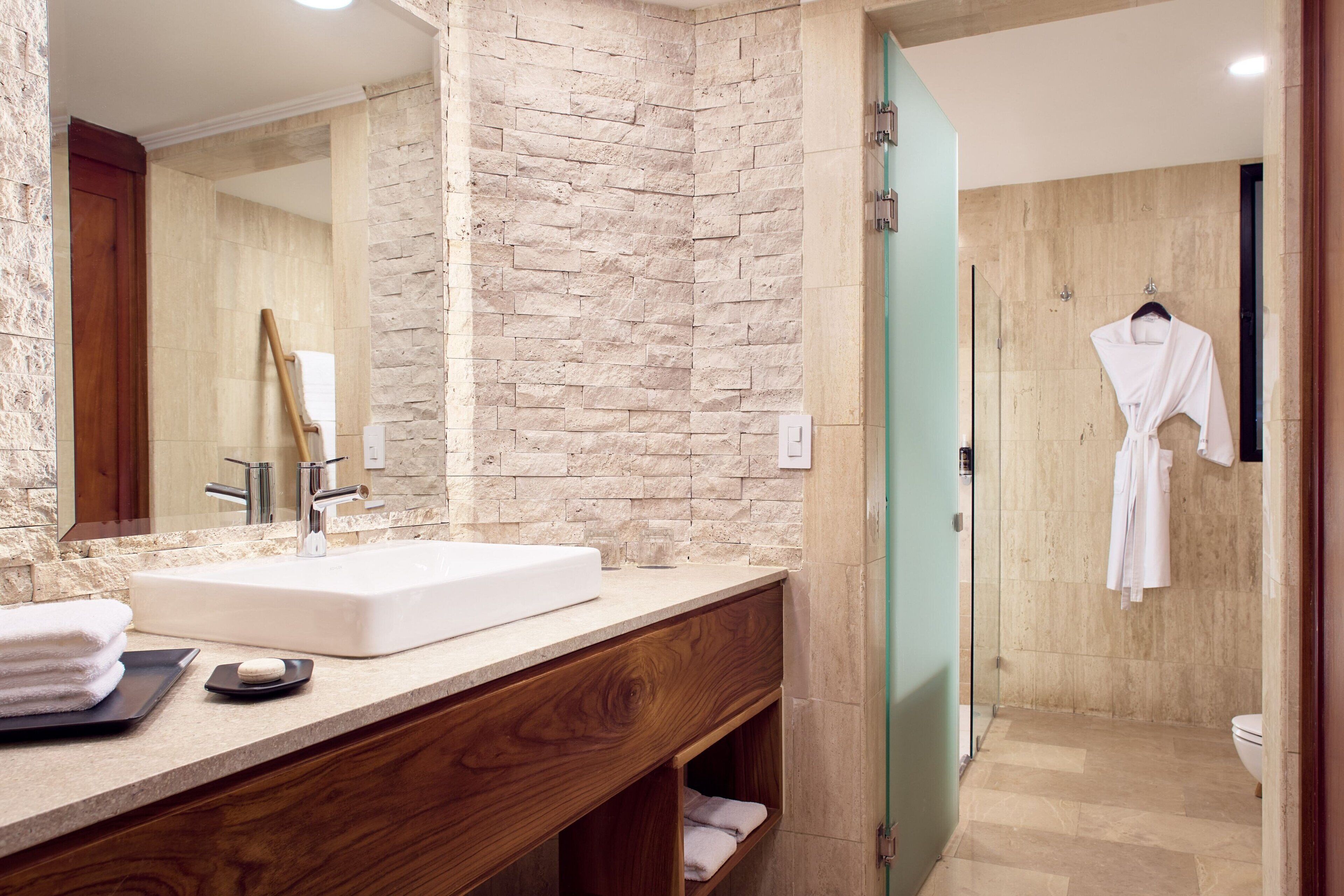 Une salle de bain moderne avec un mur en pierre, un meuble-lavabo en bois, un grand miroir et une cabine de douche en verre.